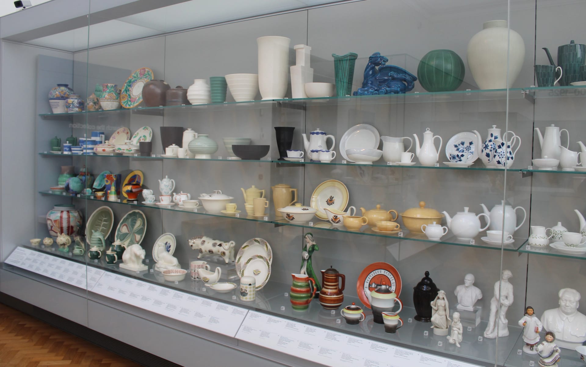 Vitrine de vidro em galeria de museu exibindo dezenas de peças de cerâmica e louça organizadas em prateleiras iluminadas.