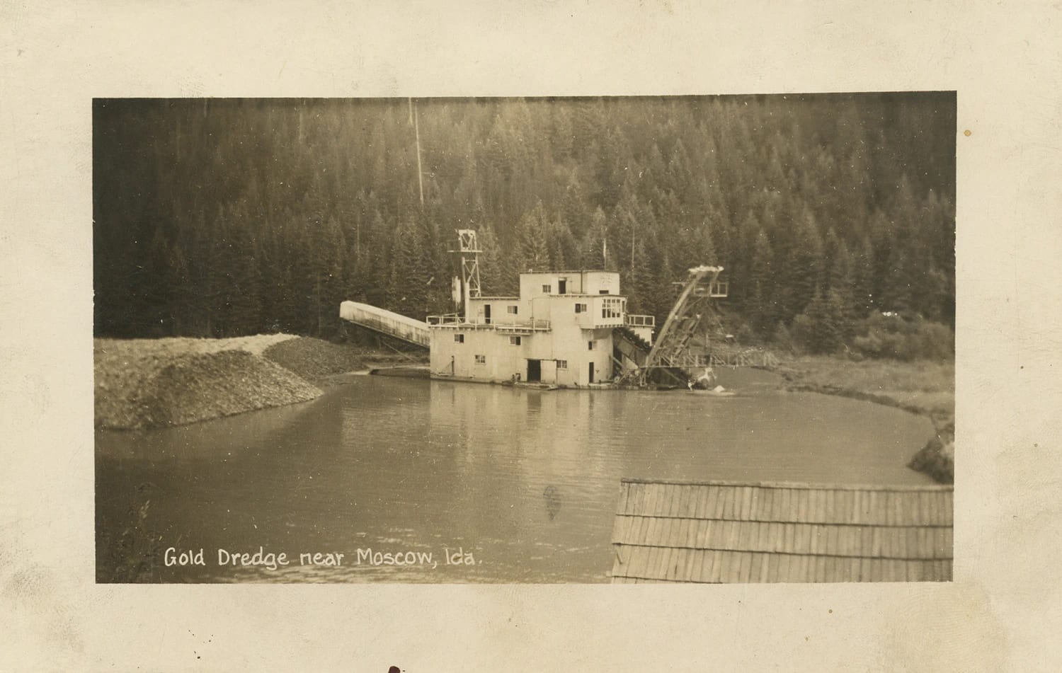Fotografia em preto e branco de uma draga de mineração de ouro flutuando em um lago cercado por floresta, Idaho, 1943.