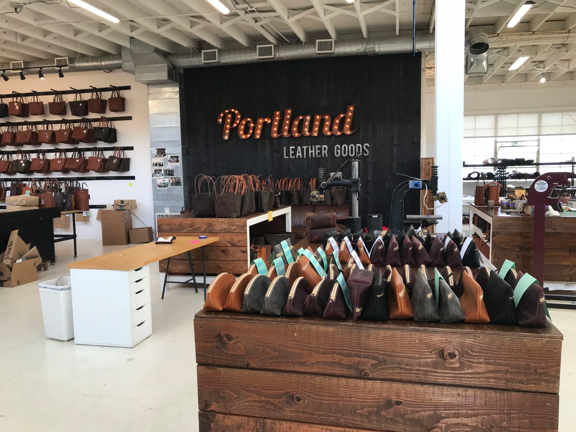 Interior de oficina de couro com bolsas artesanais expostas sobre uma bancada de madeira, diante de um letreiro luminoso com o nome 'Portland Leather Goods'.